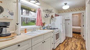 Eine kompakte Küche mit Gasherd, Spüle und weißen Schränken. Ein Fenster sorgt für natürliches Licht, und eine Tür führt in ein Schlafzimmer.