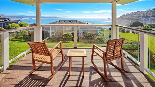 Eine überdachte Terrasse mit zwei Holzstühlen und einem Tisch, mit Blick auf eine malerische Küstenlandschaft mit Hügeln und einem Haus.