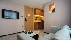 Habitación de hotel con sofá, televisor montado en la pared, una mesita, una puerta que da a una kitchenette y un cuadro en la pared.