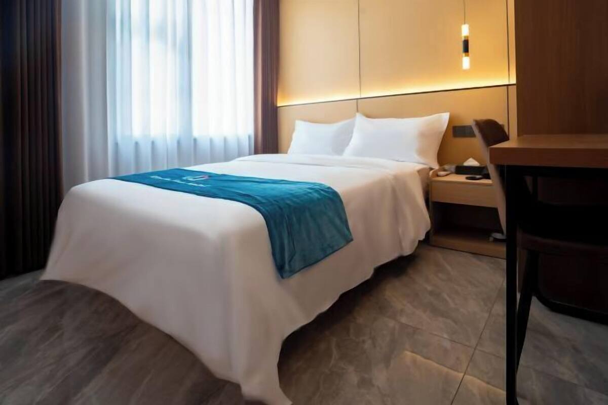 A hotel room with a bed, bedside table, lamp, and a view of the outdoors.