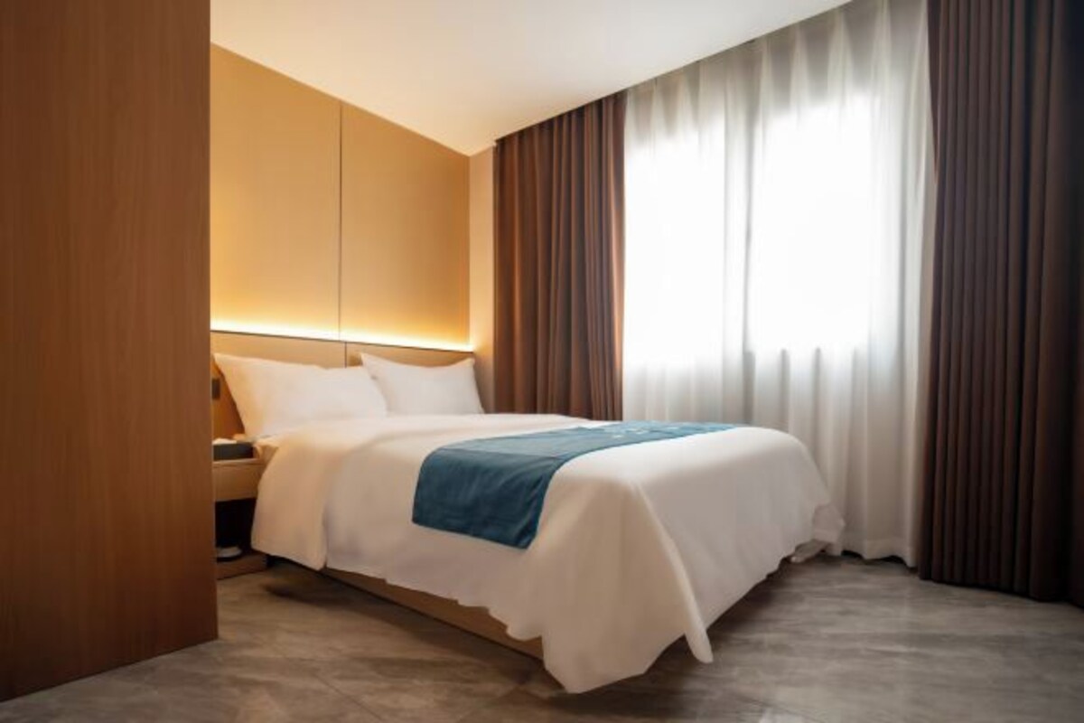 Quarto de hotel com cama, mesa de cabeceira, luminária na parede e janela com cortinas.