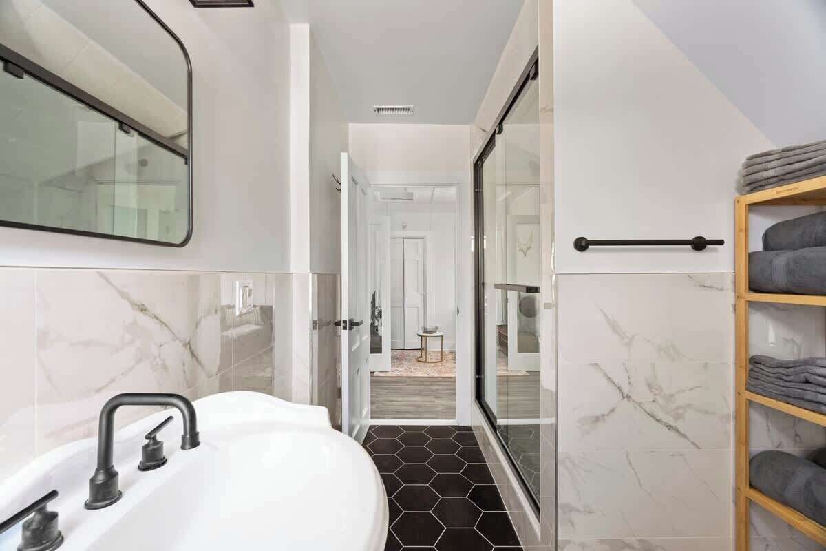 A modern bathroom with a white sink, black hexagonal tiles, a glass shower enclosure, and a wooden towel rack.