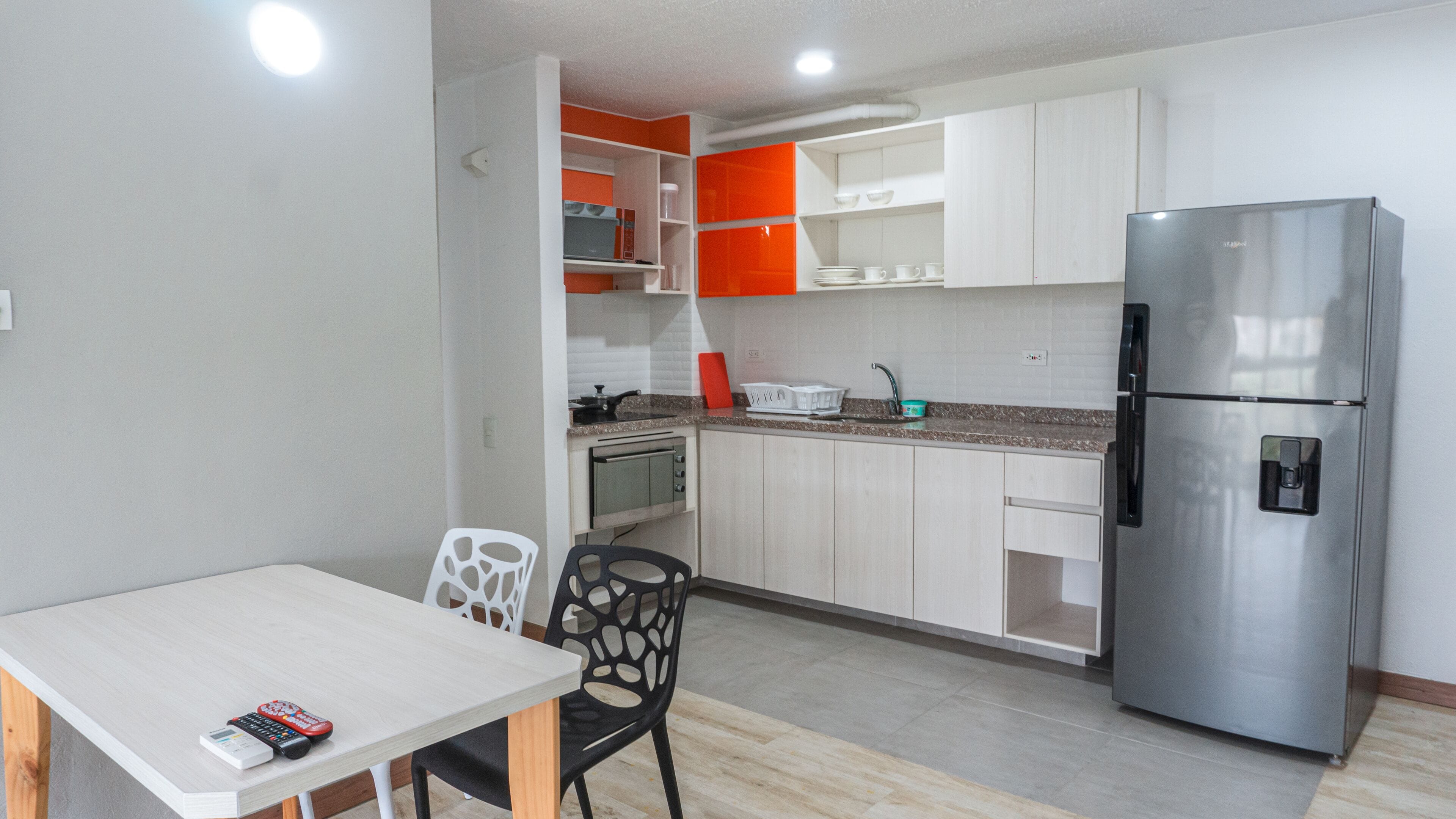 A compact kitchen with white cabinets, a stainless steel refrigerator, a microwave, and a small oven. There is a dining table with chairs and a remote control on it.