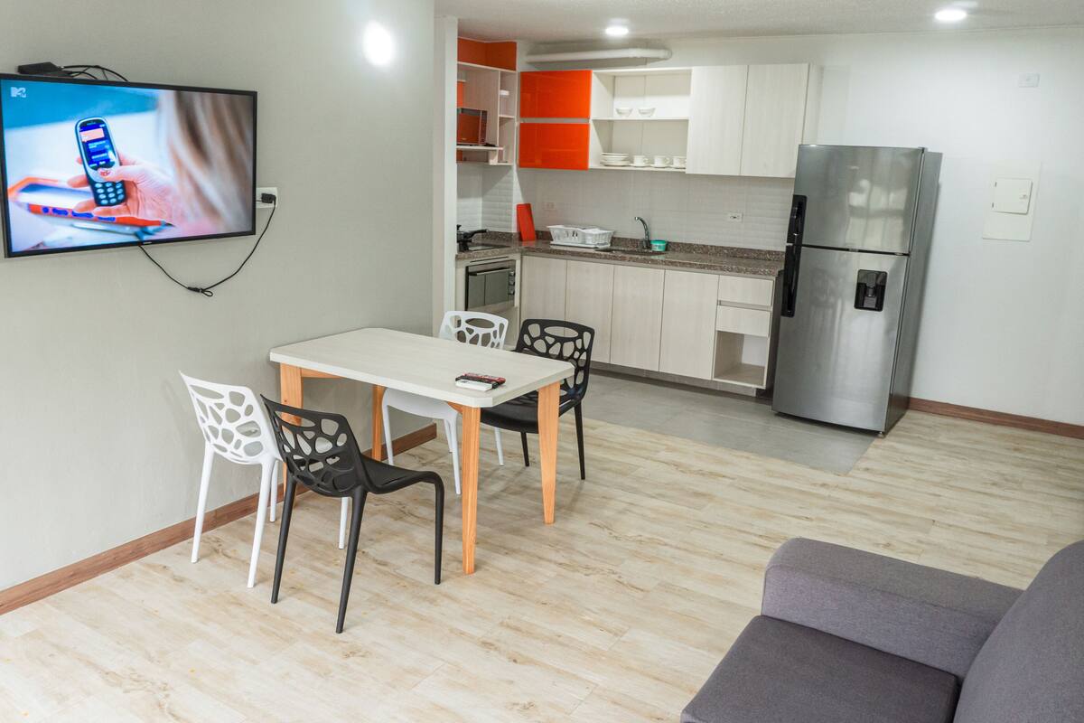 Une cuisine moderne avec des meubles blancs et orange, un réfrigérateur en acier inoxydable et un coin repas comprenant une table et des chaises blanches.