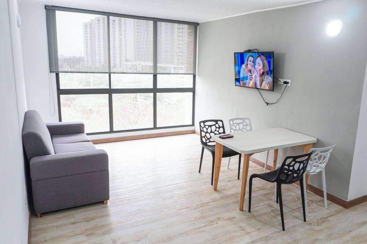 Un salon moderne avec un canapé gris, une table à manger avec des chaises, un téléviseur fixé au mur et de grandes fenêtres donnant sur des immeubles.