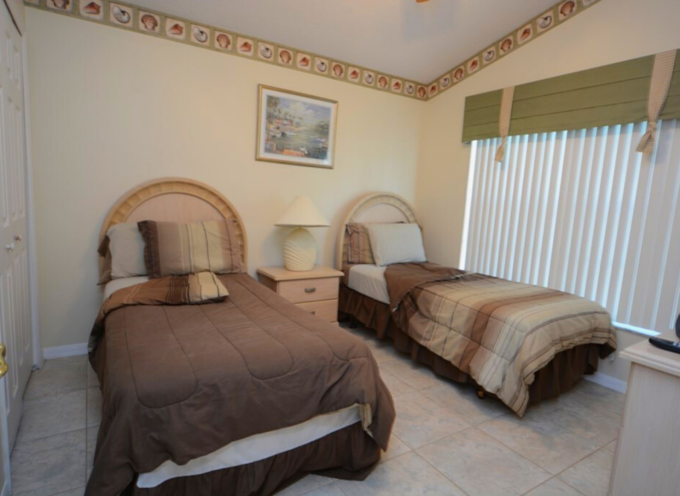 A bedroom with two beds, a nightstand, a lamp, a painting on the wall, and a window with blinds.