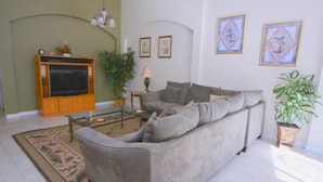 Ein Wohnzimmer mit grauem Sofa, einem Fernseher auf einem Holzstandfuß, einem Glastisch, einer Pflanze und gerahmten Bildern an der Wand.
