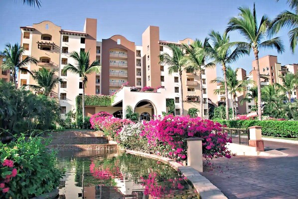 A hotel with multiple-story buildings, a central courtyard with a water feature, and lush greenery including palm trees and flowering plants.