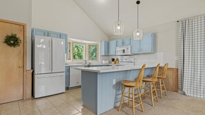 A kitchen with a central island, light blue cabinets, white appliances, and wooden bar stools. - Corolla Light Escape - Hot Tub + Beach Gear + Sunsets + Wifi (Corolla)