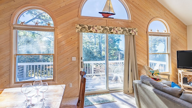 A wooden cabin interior with a dining table, chairs, a sofa, a television, and large windows with star decorations. - Corolla Light Escape - Hot Tub + Beach Gear + Sunsets + Wifi (Corolla)