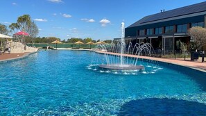 Zwembad met een fontein in het midden, omringd door ligbedden en parasols, met een gebouw op de achtergrond.