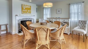 Un coin repas avec une table ronde, des chaises en osier, une cheminée et un lustre.