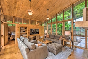 A spacious living room with a large TV, comfortable seating, and large windows offering a view of the forest.