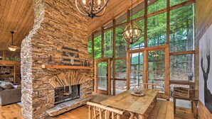 Interior rústico de una cabaña con chimenea de piedra, mesa de comedor de madera y ventanas amplias con vistas al bosque.