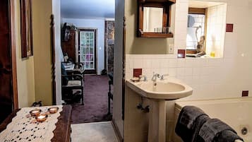 A bathroom with a sink, mirror, and bathtub. There is a door leading to another room with a chair and table.