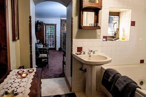 A bathroom with a sink, mirror, and bathtub. There is a door leading to another room with a chair and table. - BENTON PLACE INN (Eureka Springs)