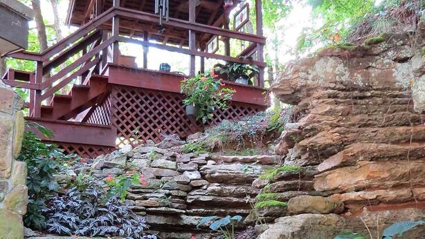 A wooden staircase leading to a wooden structure with a balcony, surrounded by lush greenery and stone walls.