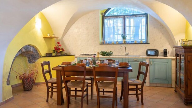 A dining area with a round table and chairs, a kitchen with a window, and a stone archway. - Apartment Giallo by Interhome (Vasia)