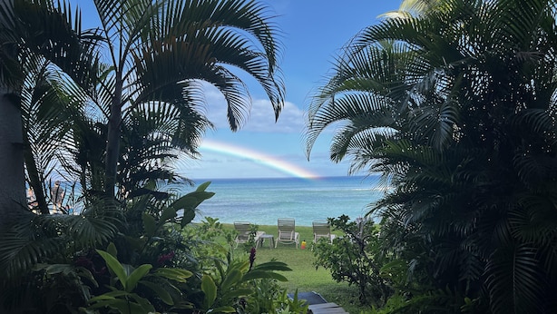 Un jardin tropical qui mène à une plage où l’on peut admirer un arc-en-ciel, une végétation luxuriante et une vue dégagée sur l’océan.