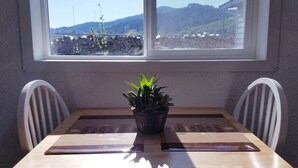 A small wooden table with two white chairs, a potted plant, and a window with a view of mountains and a stone wall. - South Sunrise Studio - Retreat in Forks (Forks)