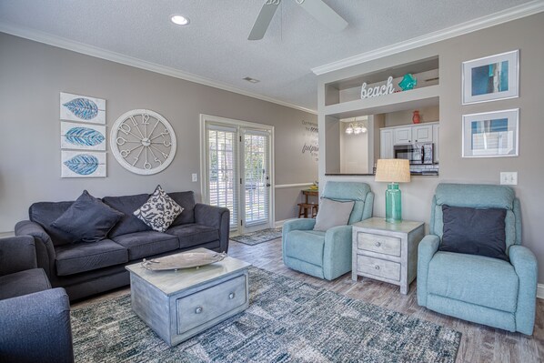 A living room with a gray sofa, teal armchairs, a coffee table, and a kitchen area in the background.