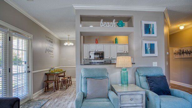 Una sala de estar con sillones azul verdosos, un comedor con muebles de madera y una cocina con alacenas blancas y un microondas.