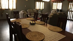 Un coin repas avec une table en bois, des sets de table en fibres tressées et des bougies. Des chaises sont disposées autour de la table, et un canapé est visible au fond.