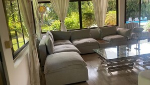 Una sala de estar moderna con un sofá seccional, una mesa de centro de vidrio y ventanas amplias con vistas a una piscina y vegetación.