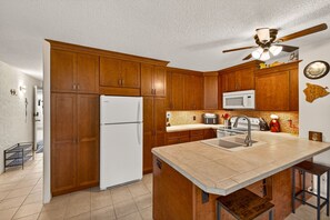Une cuisine avec des meubles de rangement en bois, un réfrigérateur blanc, un four à micro-ondes, un évier et un ventilateur de plafond.