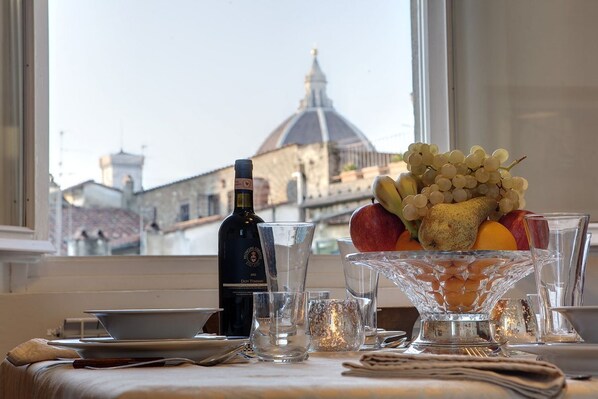 A table set with a bottle of wine, glasses, and a fruit bowl with grapes, pears, and apples.
