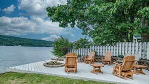 Um deck de madeira com cadeiras Adirondack e um braseiro, com vista para um lago, com uma cerca de madeira e árvores ao fundo.