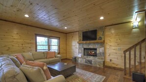 A cozy living room with a stone fireplace, wooden walls, a sofa set, and a staircase. - Luxury Mountain Cabin with Amazing Views and Hot Tub (Dog Friendly) (Blue ridge)