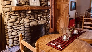 Ein Kamin aus Stein mit einem gerahmten Bild darüber, ein Holzesstisch mit einer rot-schwarzen karierten Tischdecke und ein Holzstuhl.