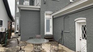 Une terrasse avec une table ronde et deux chaises, une porte blanche et un mur en briques grises.