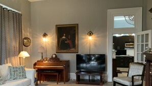 Un salon comprenant un piano, un téléviseur, un canapé et un fauteuil. Il y a un tableau au mur et deux appliques murales.