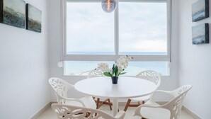 Ein Essbereich mit einem runden Tisch und weißen Stühlen, einer Orchidee im Topf sowie Meerblick durch ein großes Fenster.