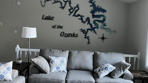 Ein Wohnzimmer mit grauem Sofa, gemusterten Kissen, einer weißen Lampe und einer Wanddekoration mit dem Schriftzug 'Lake of the Ozarks'.