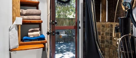 Een kleine badkamer met toilet, planken met handdoeken, een douchegedeelte en een deur naar een buitenruimte.
