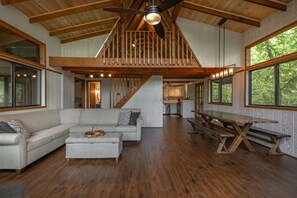 A spacious living area with a wooden ceiling, a large sofa, a dining table, and a staircase leading to an upper level.