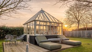 A glass conservatory with a wooden deck, seating area, and a view of the sunset. - Homestead and Black Cat Barn, Southrepps, Norfolk (Southrepps)