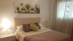 Ein ordentlich bezogenes Bett mit brauner Bettdecke und dekorativen Kissen, eine Nachttischlampe sowie ein an der Wand hängendes abstraktes Gemälde.