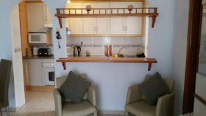Ein kompakter Wohnraum mit Küchenzeile, zwei Sesseln und einem Holzregal über der Arbeitsfläche.