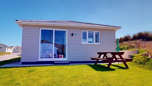 A small, single-story cottage with a sliding glass door entrance, a patio table, and a clear view of the surrounding grassy area. - G07 Tweenbanx (G7), Riviere Towans (Hayle)