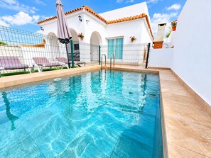 Piscina com água cristalina, cerca e um edifício com telhado e arcos azulejados.