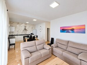 A modern living room with two sofas, a dining area with a table and chairs, a kitchen with a marble backsplash, and a wall clock.