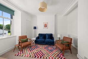 Una habitación con un sofá azul, dos sillas de madera, un tapete estampado, una lámpara y un cuadro en la pared.
