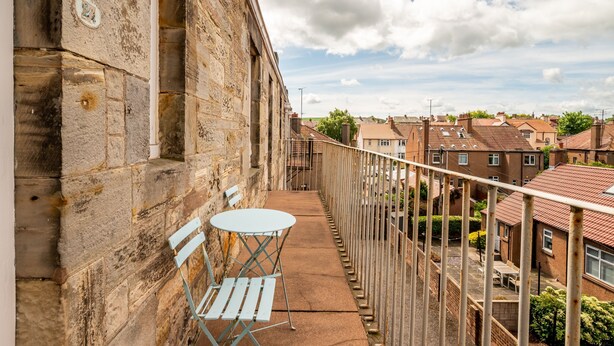 A balcony with a table and chairs, overlooking a residential area with brick houses.