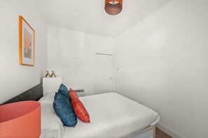 Un dormitorio con cama, almohadas, una mesita de noche, un cuadro enmarco, una puerta y una lámpara en el techo.