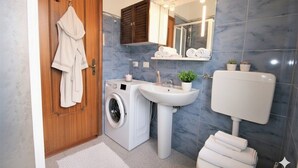 Un baño moderno con puerta de madera, lavadora, lavabo, inodoro y espejo con luces.