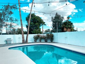 A swimming pool with a white tiled edge, surrounded by a white wall, under string lights, with trees and a clear sky in the background.